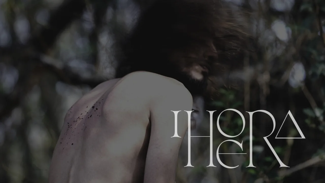 A man seen from behind, bare-shouldered, his hair in motion against blurred trees, the Ihoera logotype across the frame